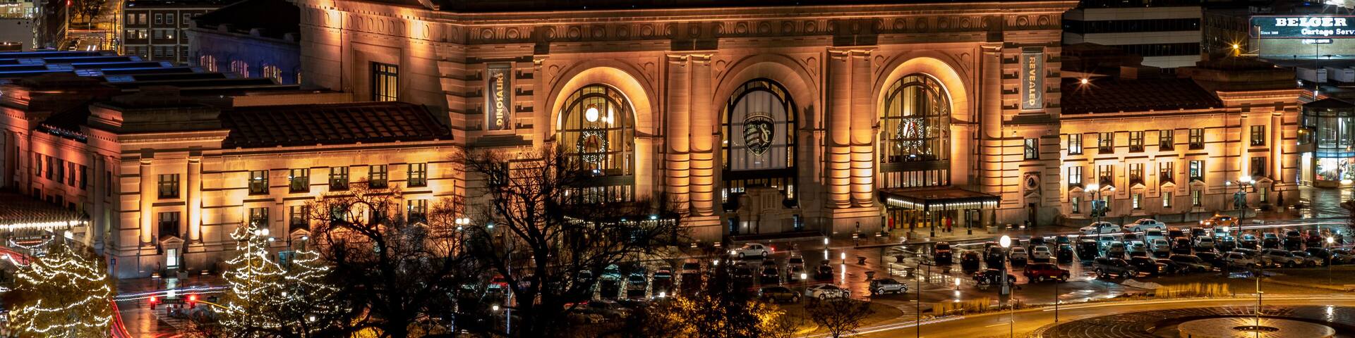 Un horizonte urbano nocturno con edificios iluminados, un edificio histórico destacado con ventanas arqueadas y un cruce de calles concurrido con automóviles.