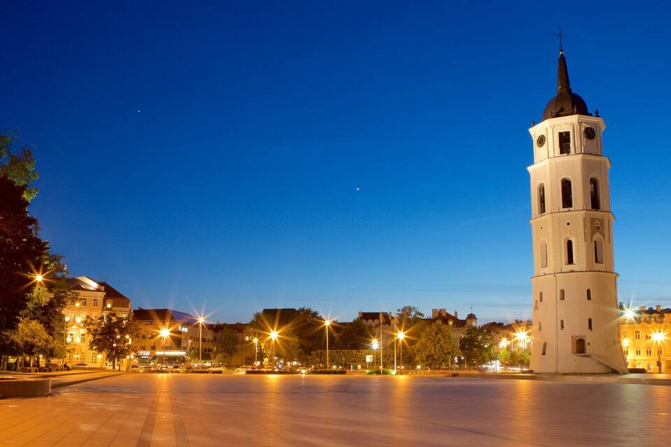 A historic tower with a clock, surrounded by a plaza with trees and street lamps.