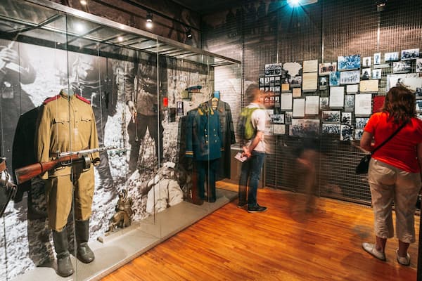 En udstilling på et museum med dukker i militæruniformer, et gevær og et baggrundsbillede af historiske fotografier.