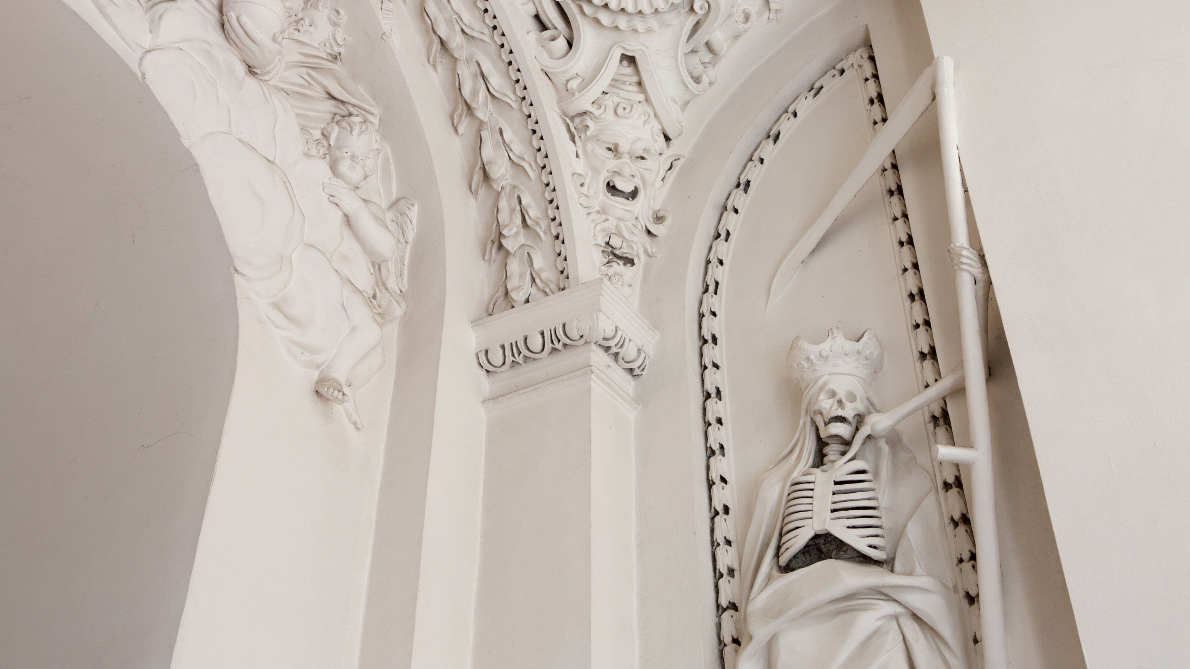 Une sculpture architecturale blanche et détaillée, représentant une figure squelettique, ornée de sculptures complexes et surmontée d’une couronne.