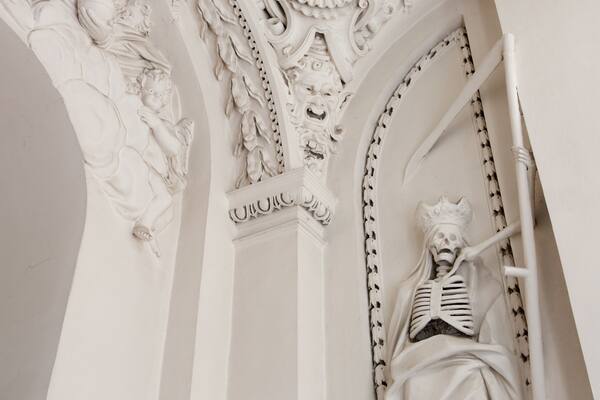 Une sculpture architecturale blanche et détaillée, représentant une figure squelettique, ornée de sculptures complexes et surmontée d’une couronne.