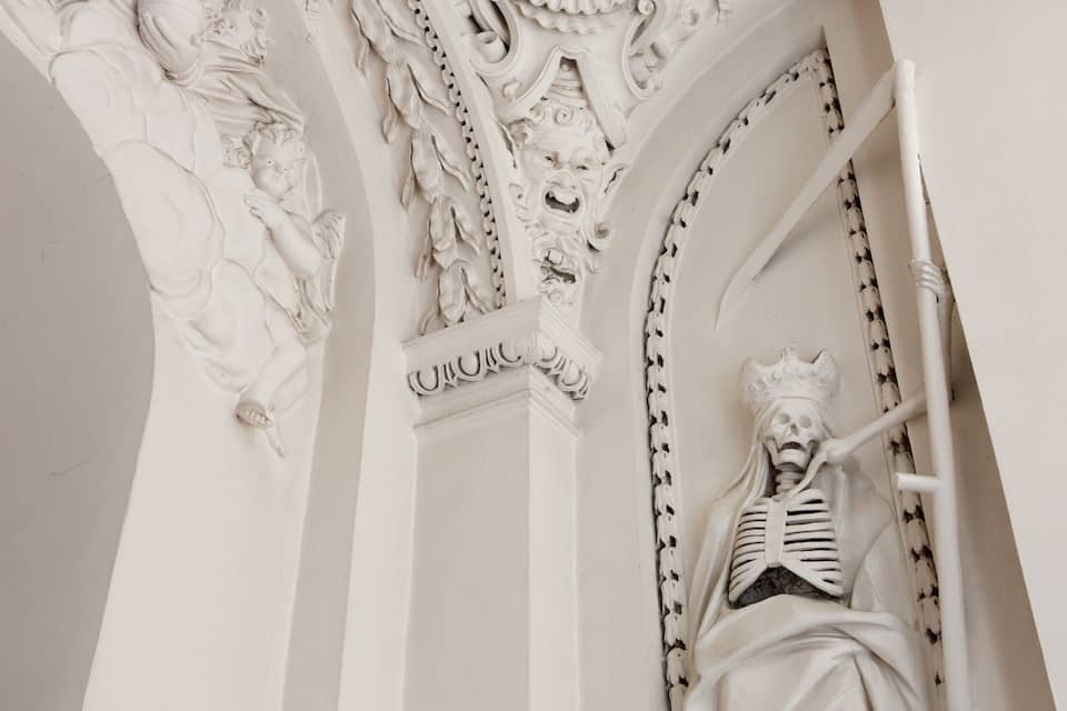Une sculpture architecturale blanche et détaillée, représentant une figure squelettique, ornée de sculptures complexes et surmontée d’une couronne.