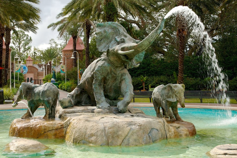 A bronze elephant fountain with water spouting from its trunk, surrounded by palm trees and a brick building in the background.
