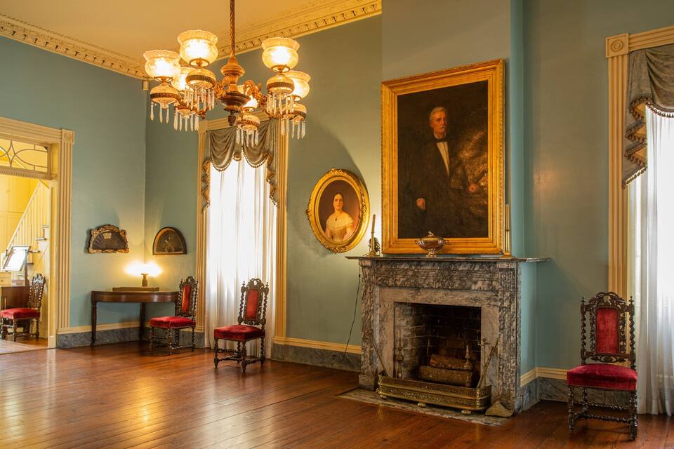 Um salão imponente com lareira de mármore, cadeiras ornamentadas, um retrato grande e um candelabro.