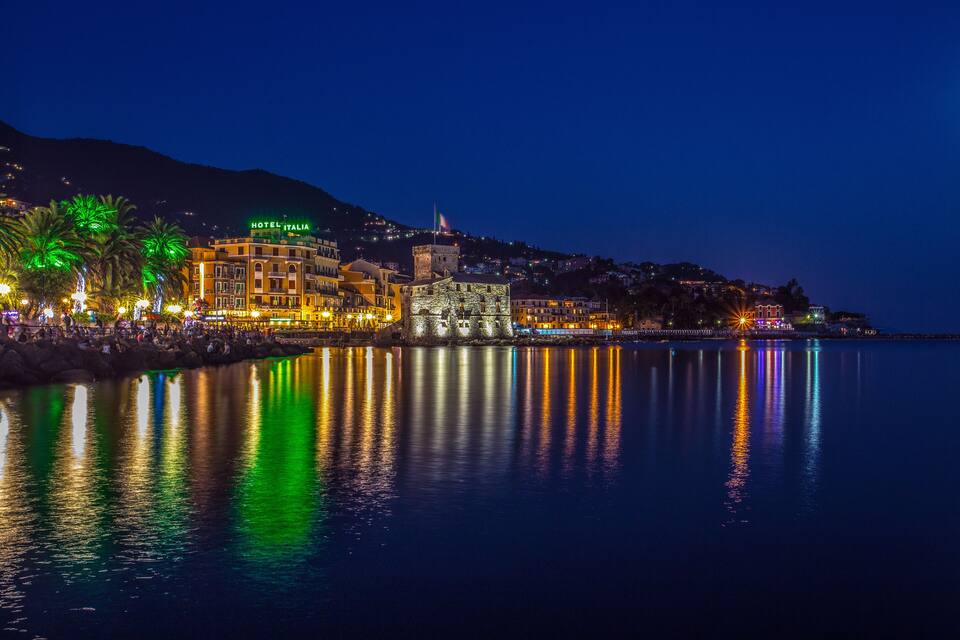 A coastal town at night with illuminated buildings, a prominent hotel, and a calm sea reflecting the lights.