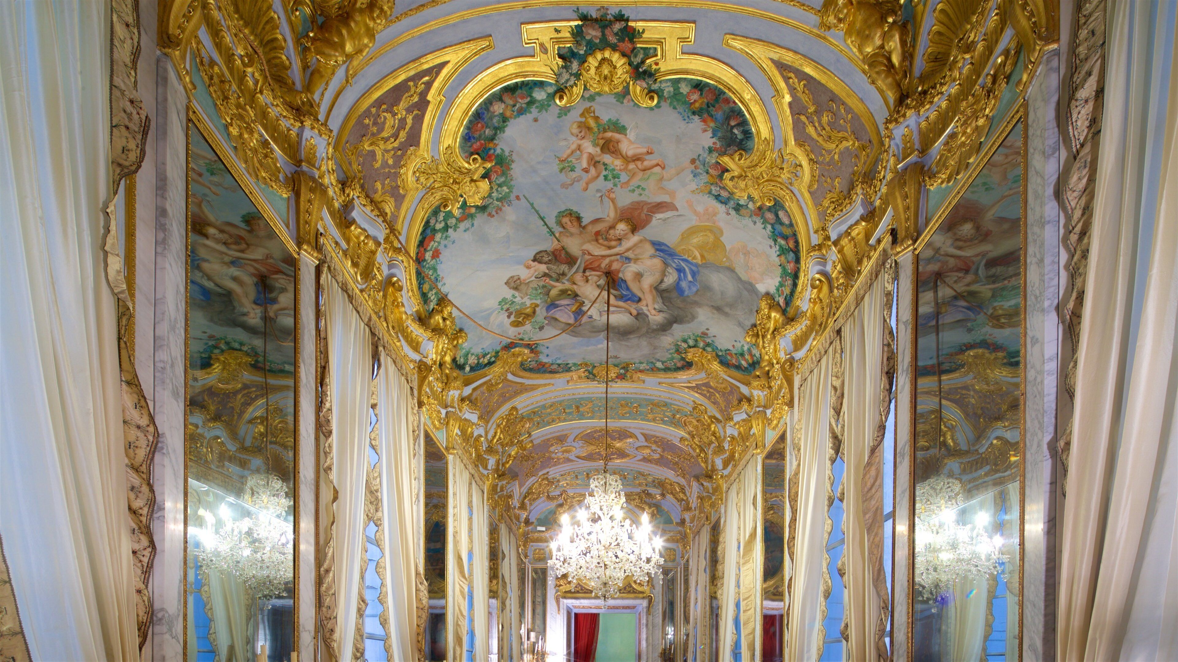 An ornate hallway with gold decorations, a chandelier, and a fresco on the ceiling.