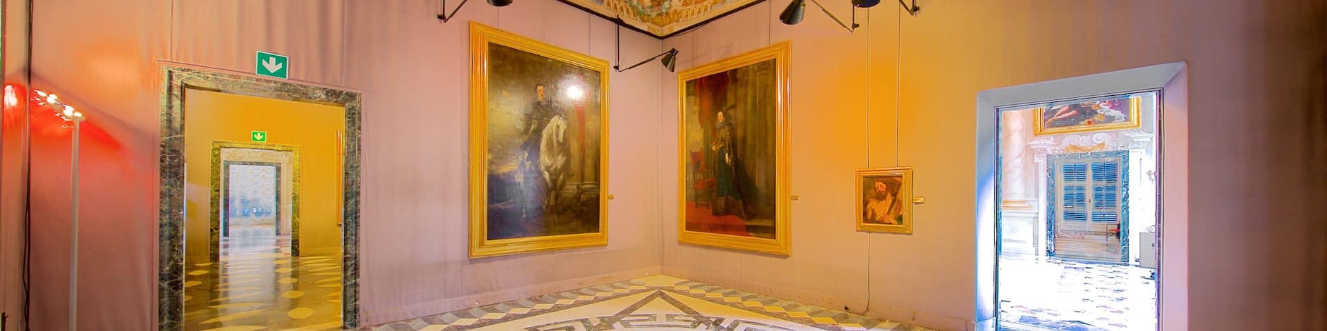 A grand hallway with ornate ceiling frescoes, large framed paintings, and a patterned floor.