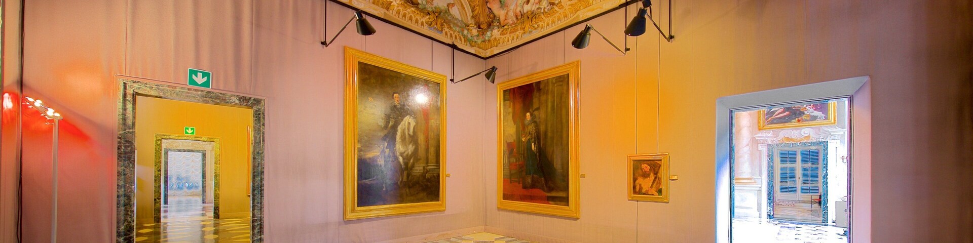 A grand hallway with ornate ceiling frescoes, large framed paintings, and a patterned floor.