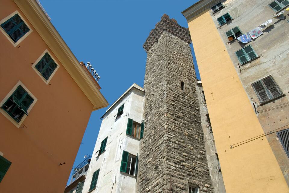 Una torre de pedra alta, un edifici amb persianes verdes i un edifici amb façana groga.