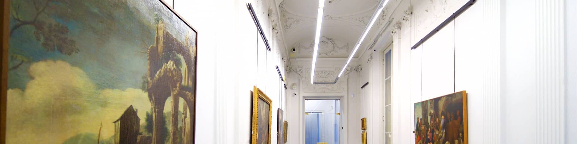 A hallway with classical architecture, featuring a black and white patterned floor, white walls, and large paintings.