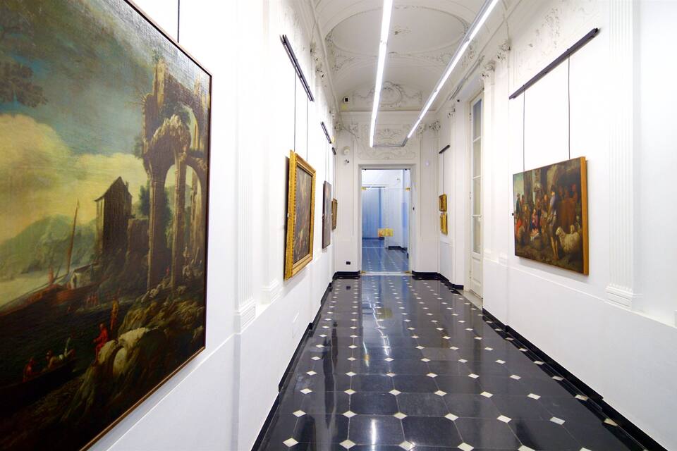 Un couloir à l’architecture classique, avec un sol à motifs en noir et blanc, des murs blancs et de grandes peintures.