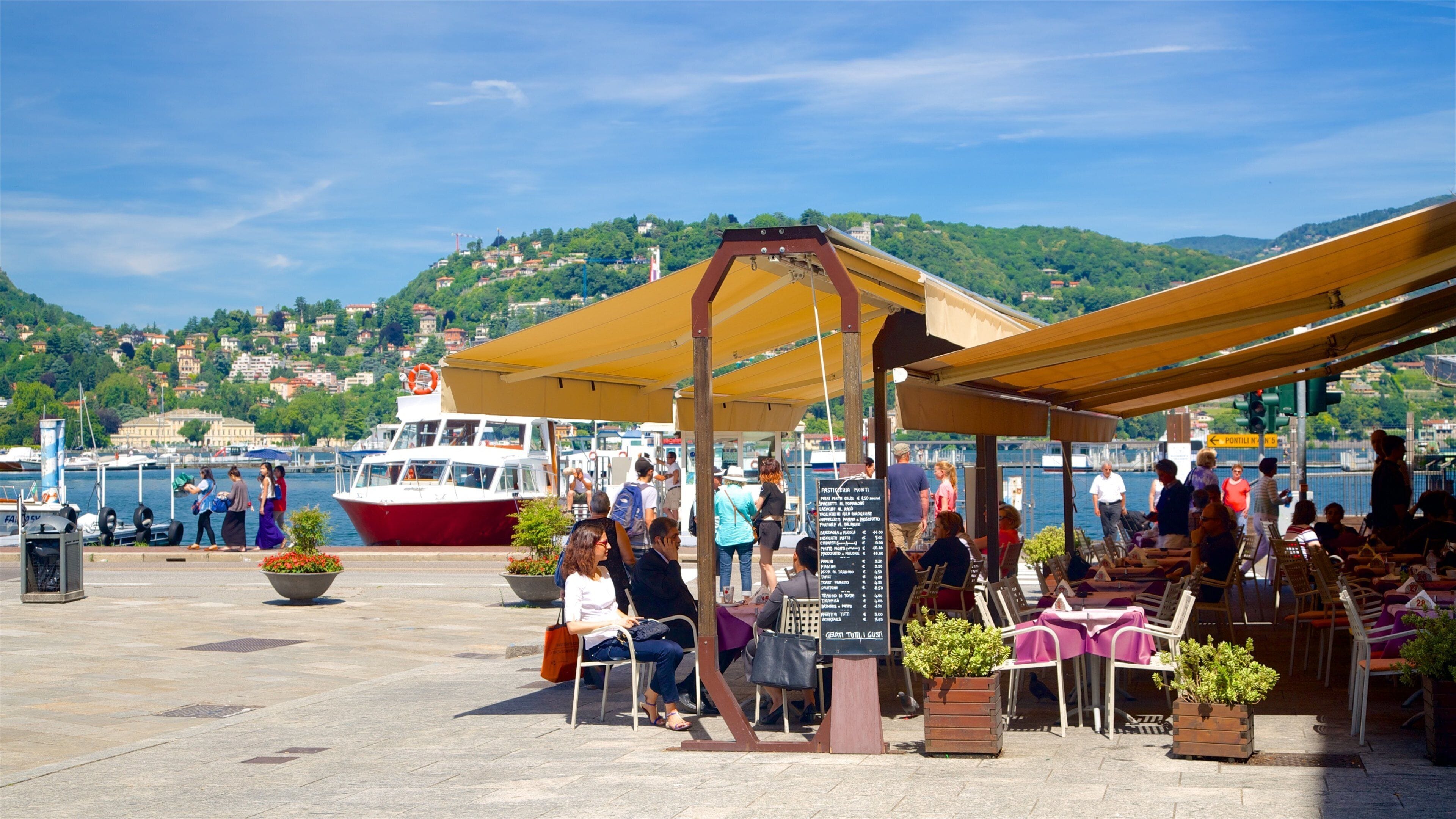 A waterfront café with outdoor seating, a red boat docked nearby, and a hillside town in the background.