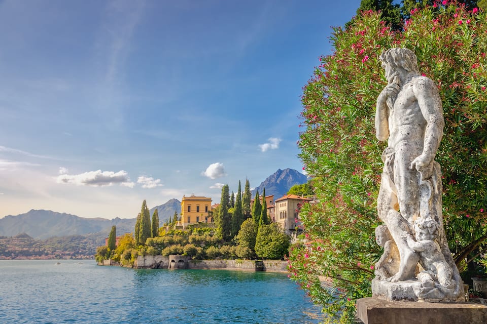 Una estatua junto a un lago, con edificios y montañas al fondo.