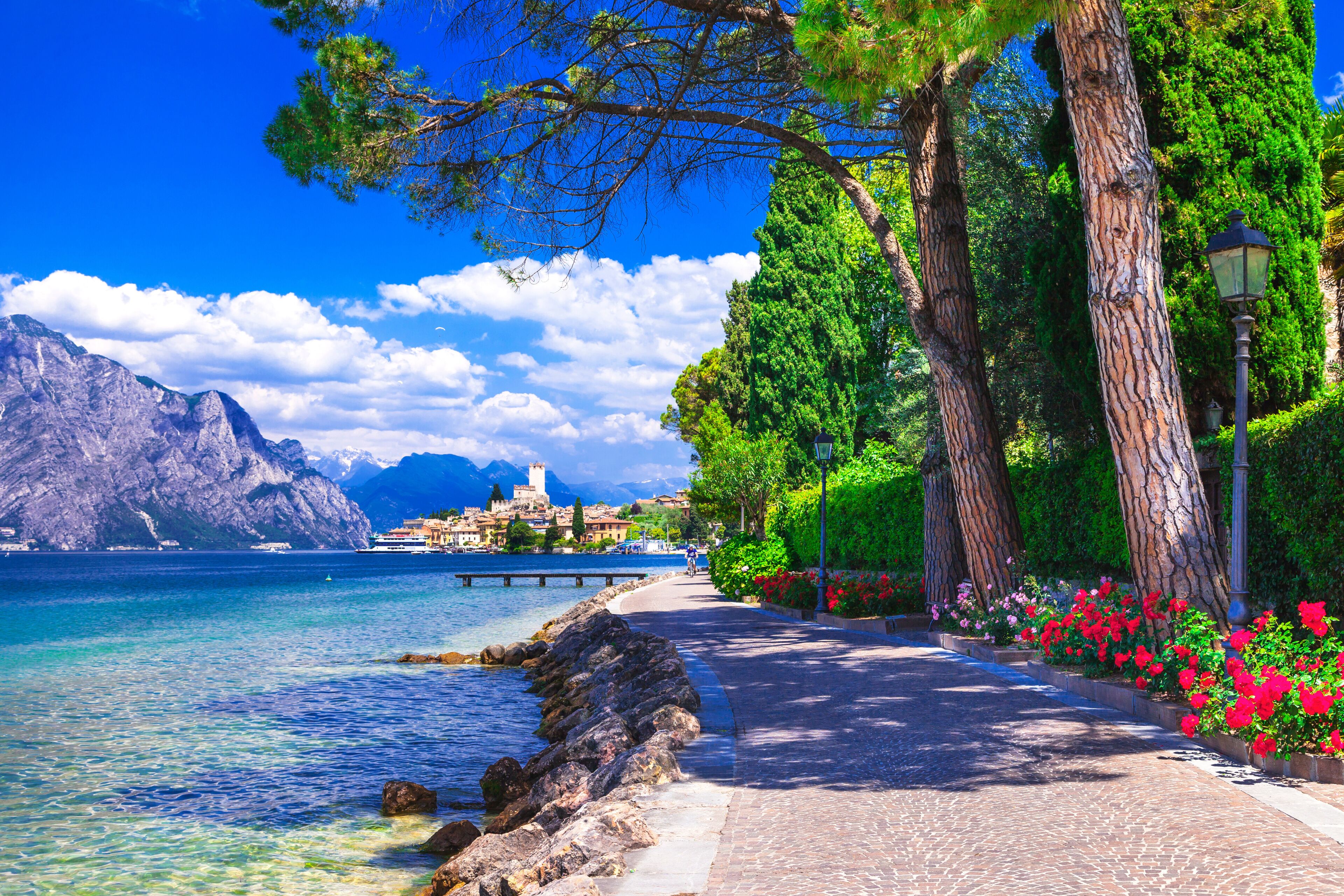 Scenic lakeside promenade in Malcesine, Lake Garda. Sunny walking path under pine trees with a view of the historic town and Scaliger Castle.