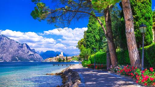 Scenic lakeside promenade in Malcesine, Lake Garda. Sunny walking path under pine trees with a view of the historic town and Scaliger Castle.