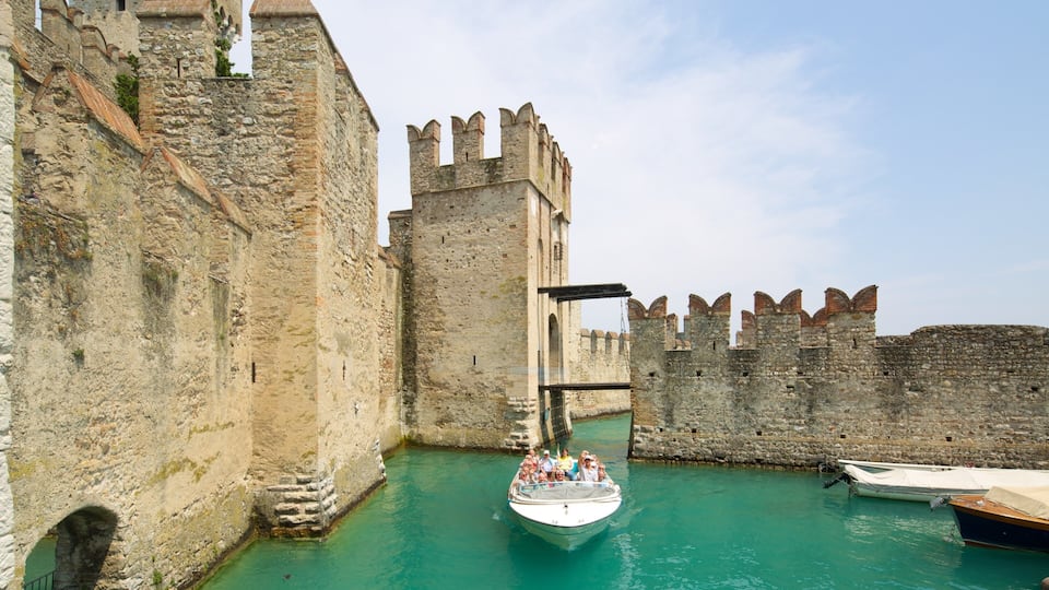 A historic stone wall with a tower, a boat with passengers, and a clear blue water canal.