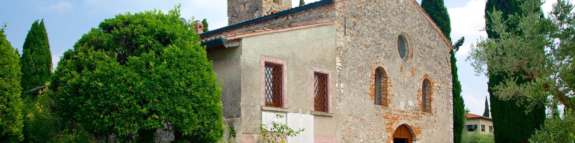 Un bâtiment historique en pierre, doté d’un clocher, entouré d’une végétation luxuriante et de grands cyprès.