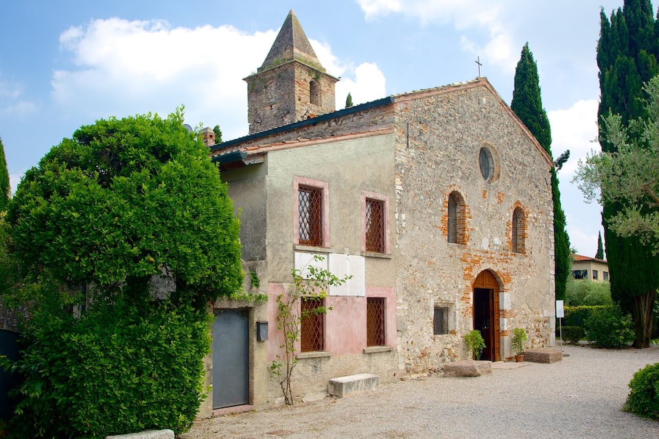 Un bâtiment historique en pierre, doté d’un clocher, entouré d’une végétation luxuriante et de grands cyprès.