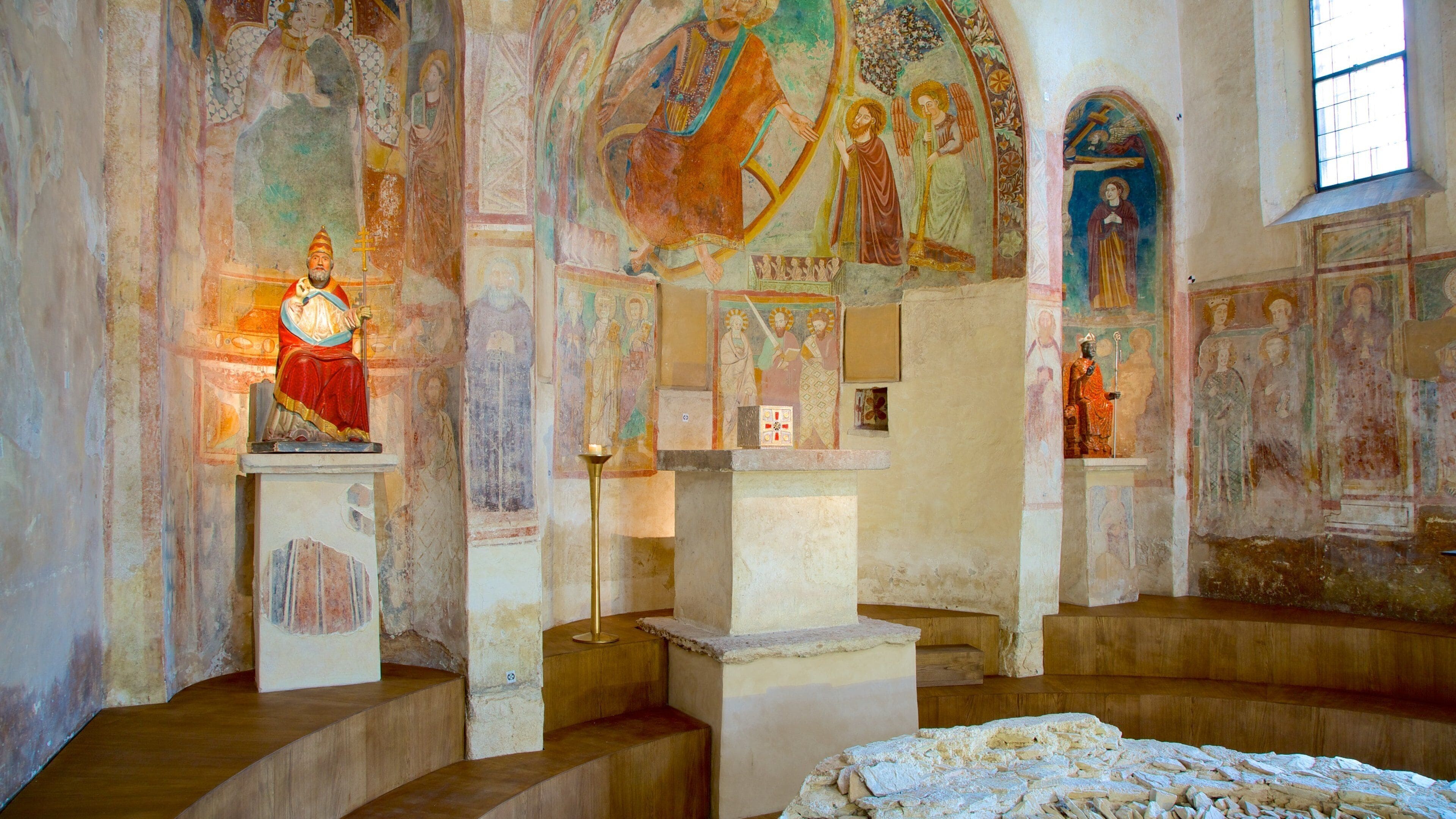 An interior with religious frescoes, a central figure on a pedestal, and a cross.