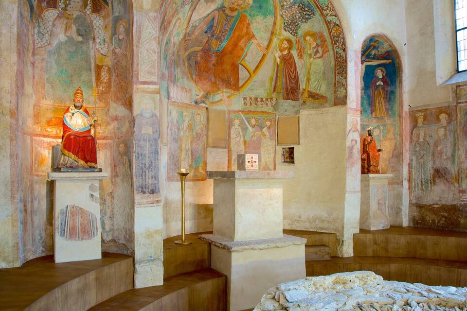 An interior with religious frescoes, a central figure on a pedestal, and a cross.