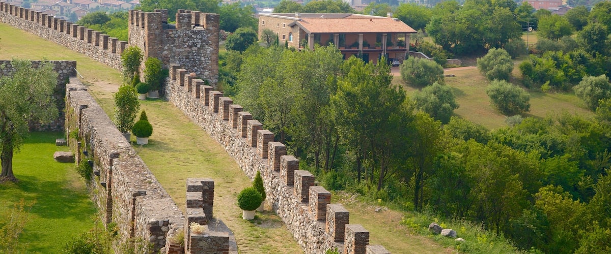 Una antigua muralla de piedra con una torre imponente, rodeada de abundante vegetación y un pueblo lejano.