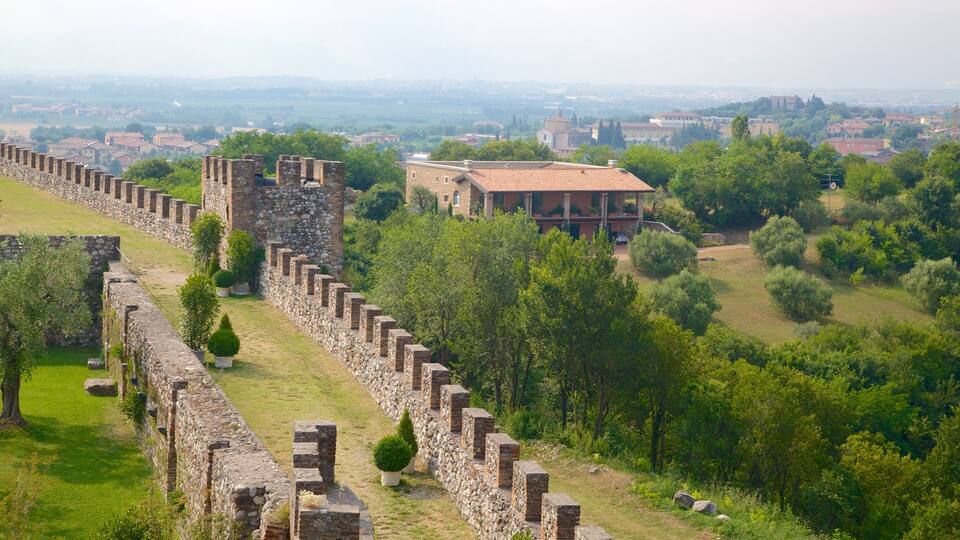 A historic stone wall with a large tower, surrounded by lush greenery and a distant town.