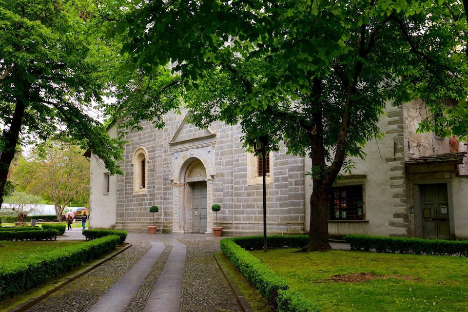 Un bâtiment historique en pierre, doté de fenêtres en arc de cercle et d’une porte d’entrée centrale, entouré de haies bien entretenues et d’un chemin pavé.