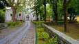 En stenhævet sti fører til en kirke med buede vinduer og en dør, omgivet af træer og en stenvæg.