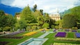 A well-manicured garden with various flower beds, a fountain, and a large building with a spire in the background.