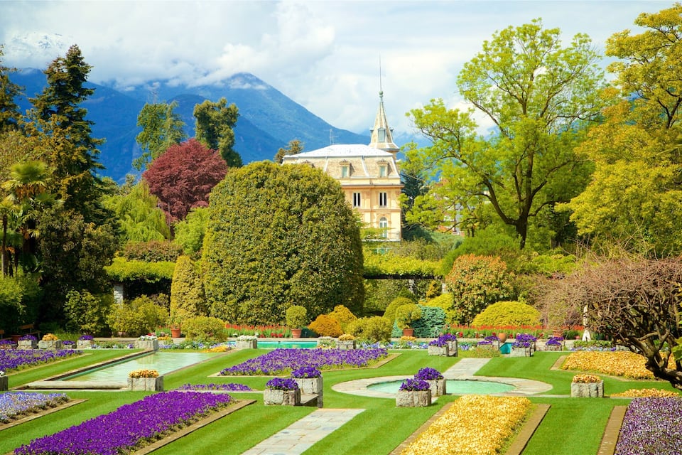 Un jardin bien entretenu, agrémenté de différents parterres de fleurs, une fontaine centrale et un bâtiment surmonté d’une flèche, visible au loin.