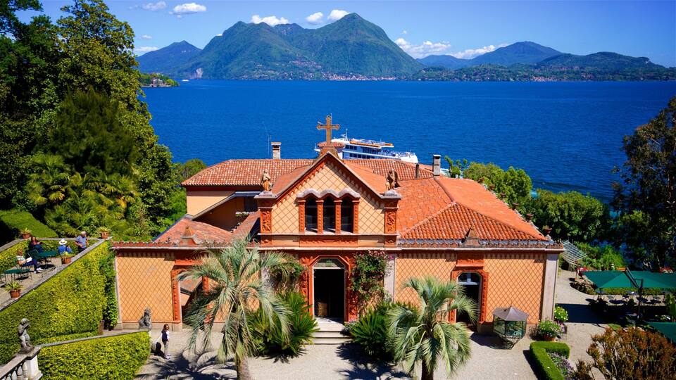A church with a red roof and a cross, surrounded by lush greenery and palm trees, with a lake and mountains in the background.