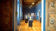 A museum corridor with wooden floors, blue walls, and classical ceiling artwork.