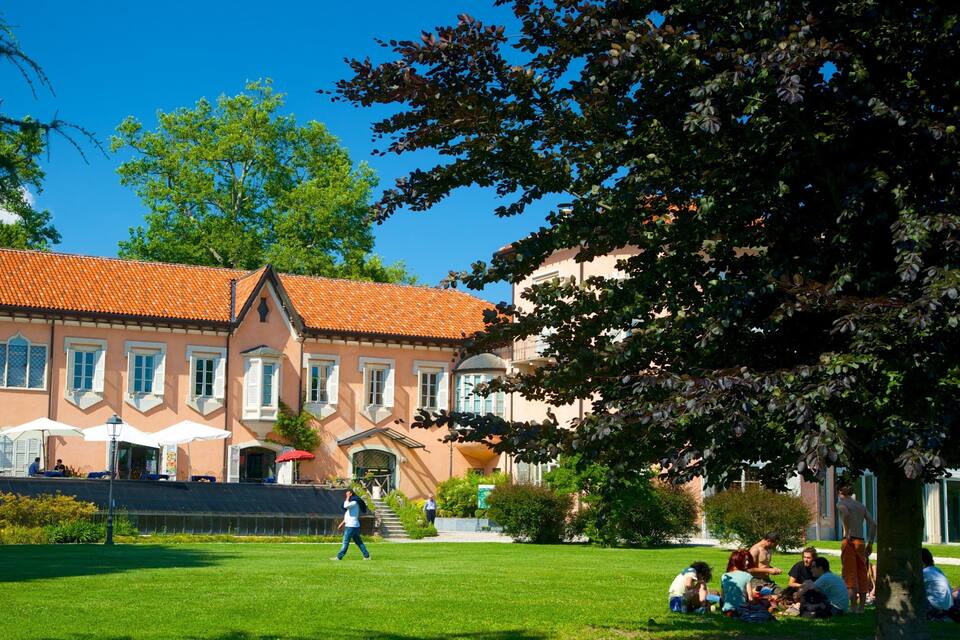 A historic building with a red-tiled roof and white shutters, surrounded by a well-maintained lawn and trees.