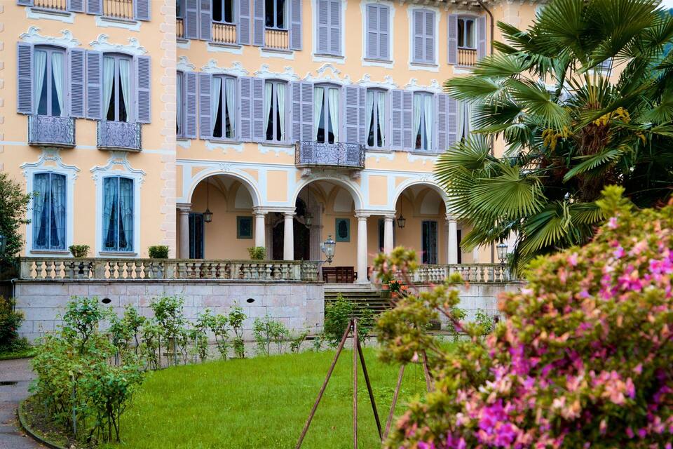 Un edifici gran i ornamentat amb diverses finestres i balcons, envoltat de vegetació exuberant i un jardí ben cuidat.