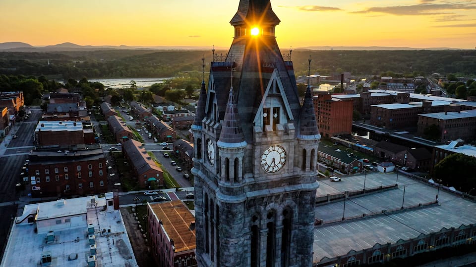 A clock tower with a spire, surrounded by buildings and a river in the background.