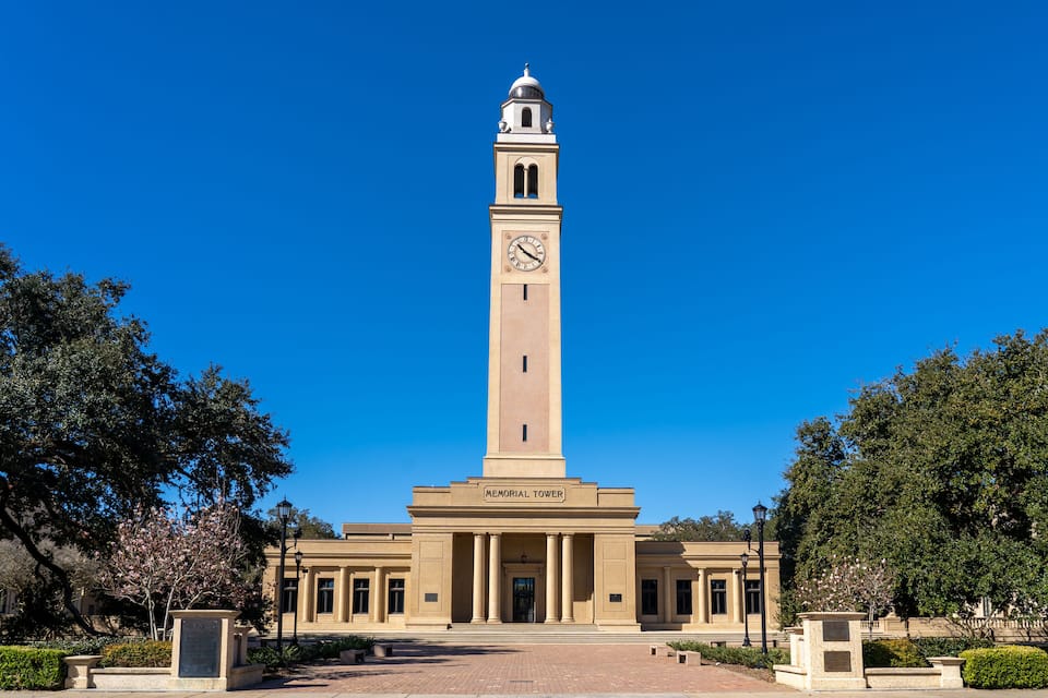 Uma torre Memorial alta, bege, com um relógio, rodeada por árvores e um caminho pavimentado.