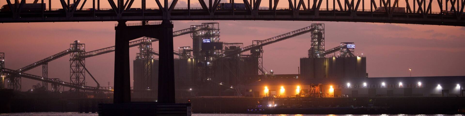 A large steel bridge spanning a body of water at dusk with industrial structures in the background.