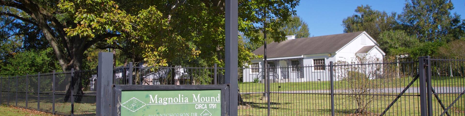 A green sign with white lettering reading "Magnolia Mound Circa 1791" and "2161 Nichols Dr." in front of a white house with a black fence and trees in the background.