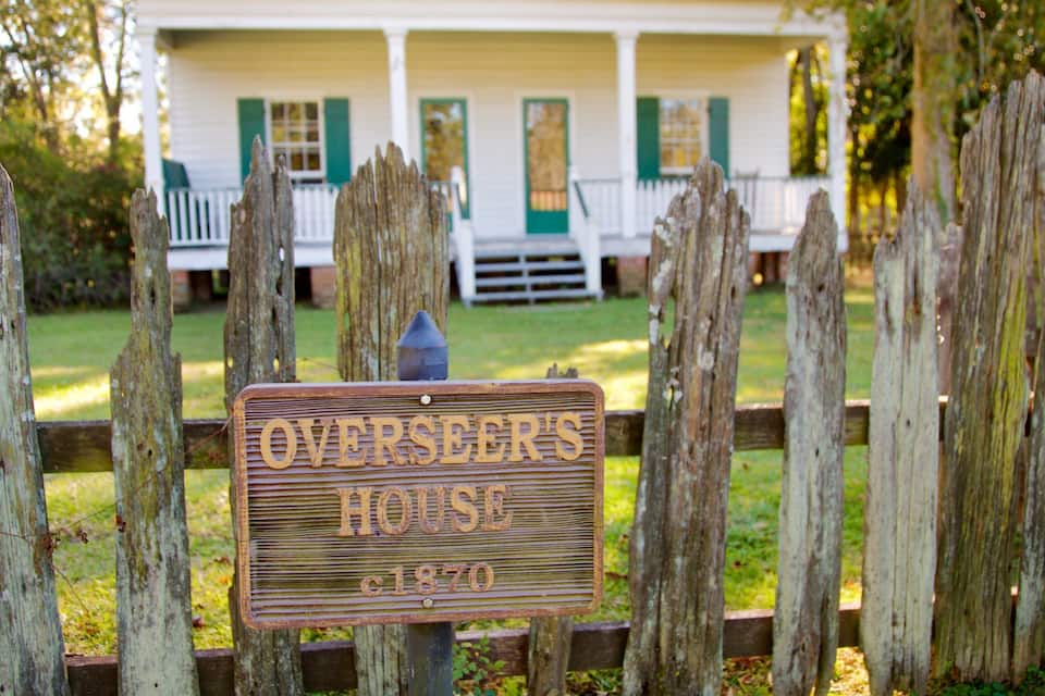A wooden sign with the text "Overseer's House c1870" in front of a white house with green shutters.