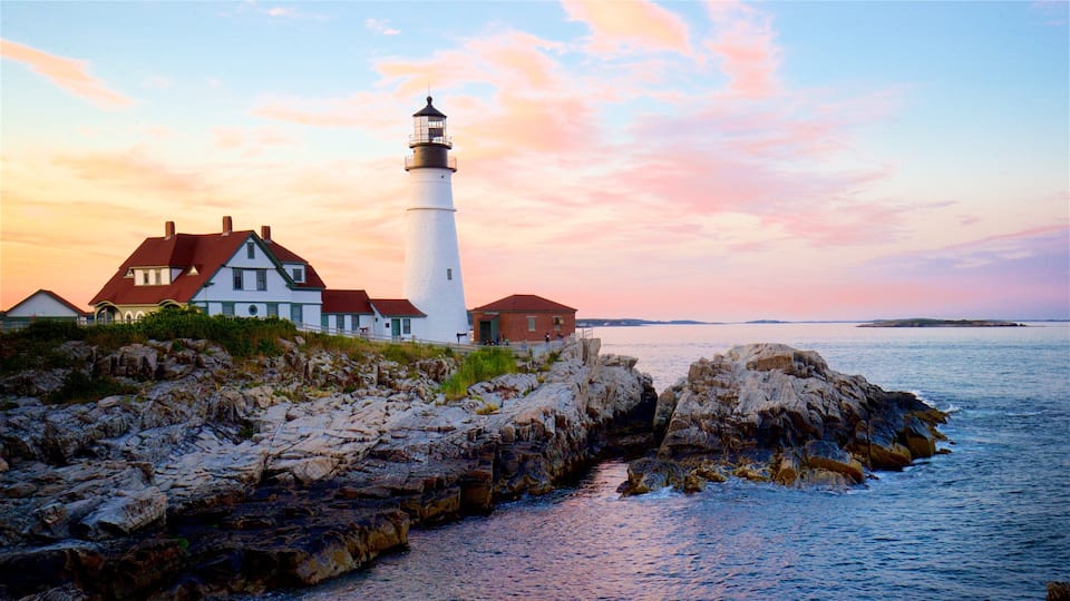A lighthouse, a white house with a red roof, and rocky coastline at sunset.