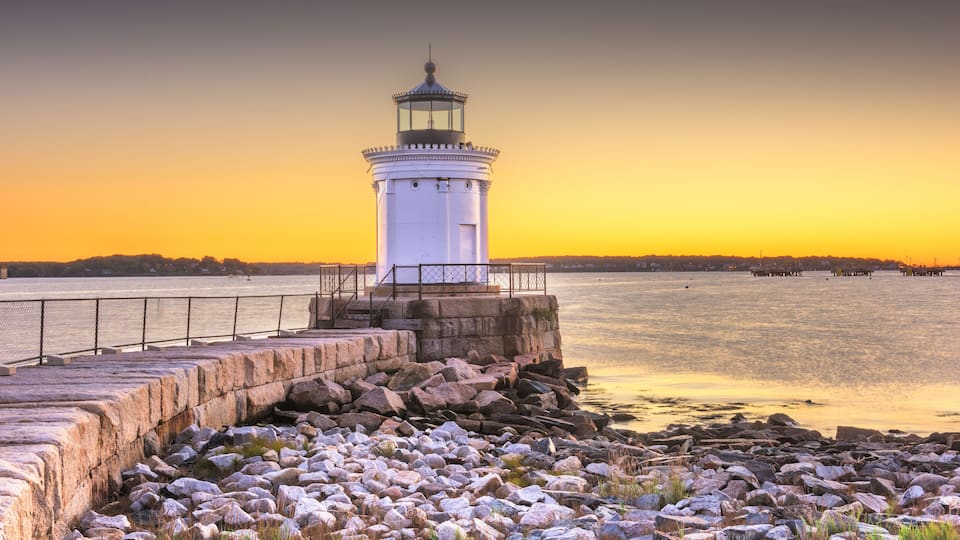 A white lighthouse on a rocky shore with a calm sea and a sunset in the background.