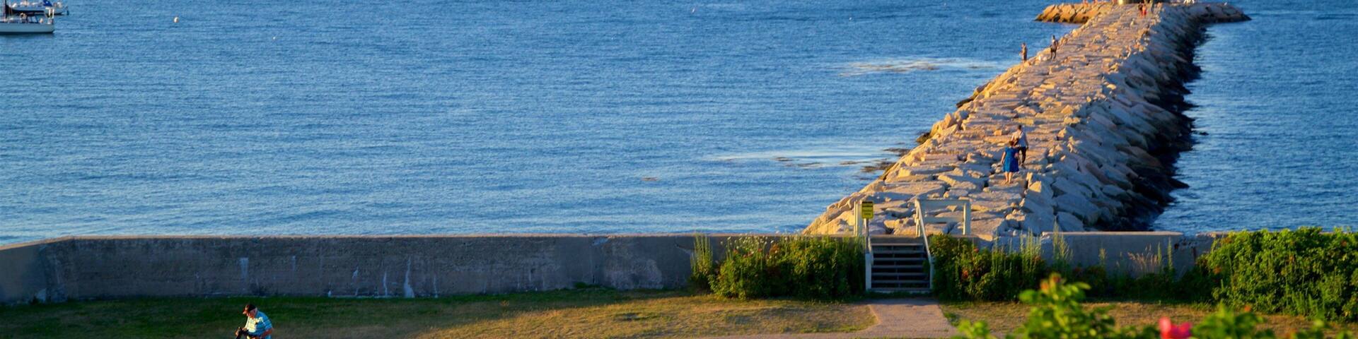 A lighthouse on a breakwater, a grassy area with a path, and calm water with boats.