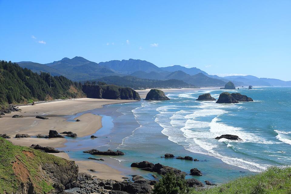 Un paisaje costero con playa arenosa, afloramientos rocosos y una cadena montañosa al fondo.