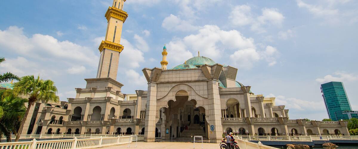 A large mosque with a tall minaret and a dome, surrounded by a paved area and palm trees.