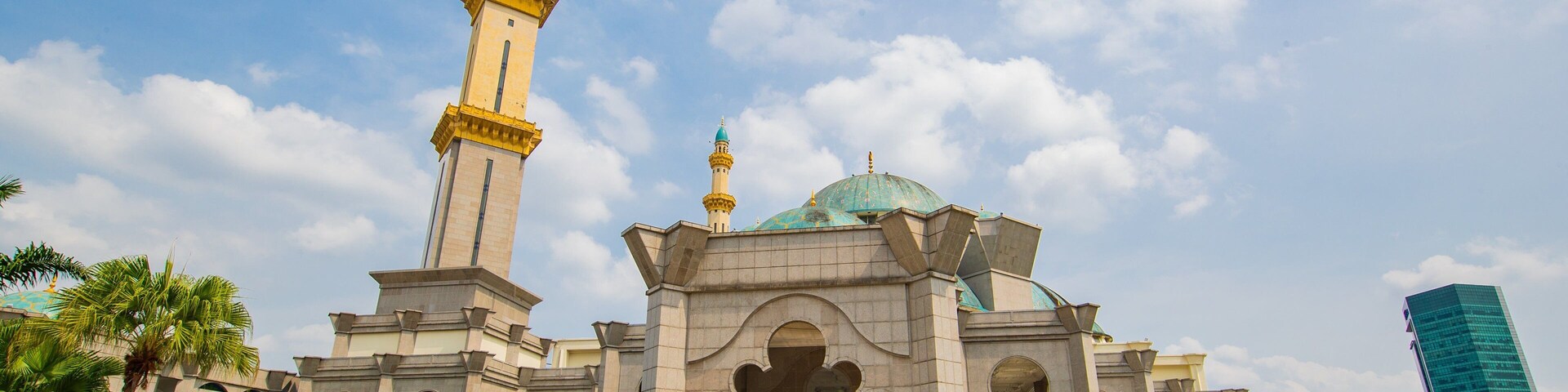 A large mosque with a tall minaret and a dome, surrounded by a paved area and palm trees.