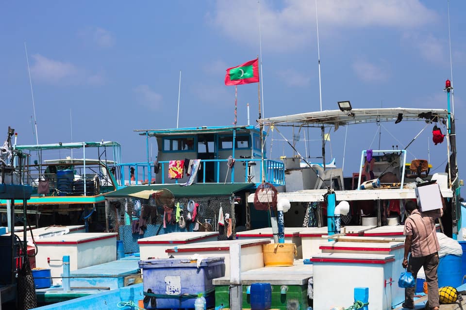 Un port animat amb diverses embarcacions amarrades, una bandera destacada i un home portant una caixa.