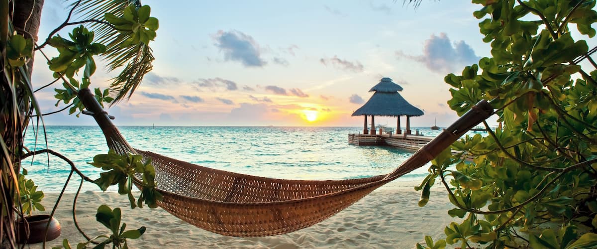A hammock on a sandy beach with a thatched-roof structure in the background and palm trees framing the scene.