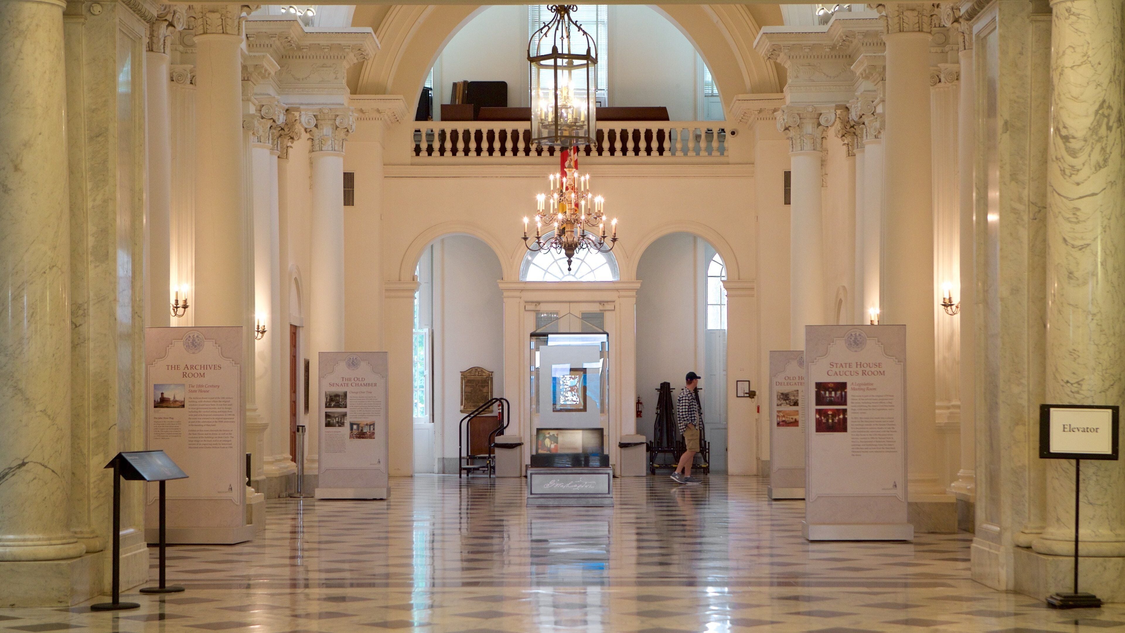 Een statige zaal met marmeren zuilen, een kroonluchter en informatiepanelen over historische kamers.
