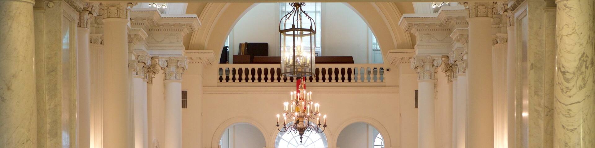 A grand hall with marble columns, a chandelier, and informational displays about historical rooms.