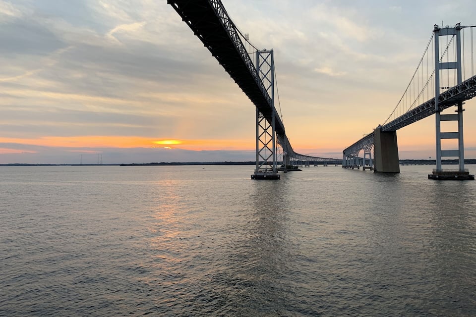 A large suspension bridge over a calm body of water with a sunset in the background.